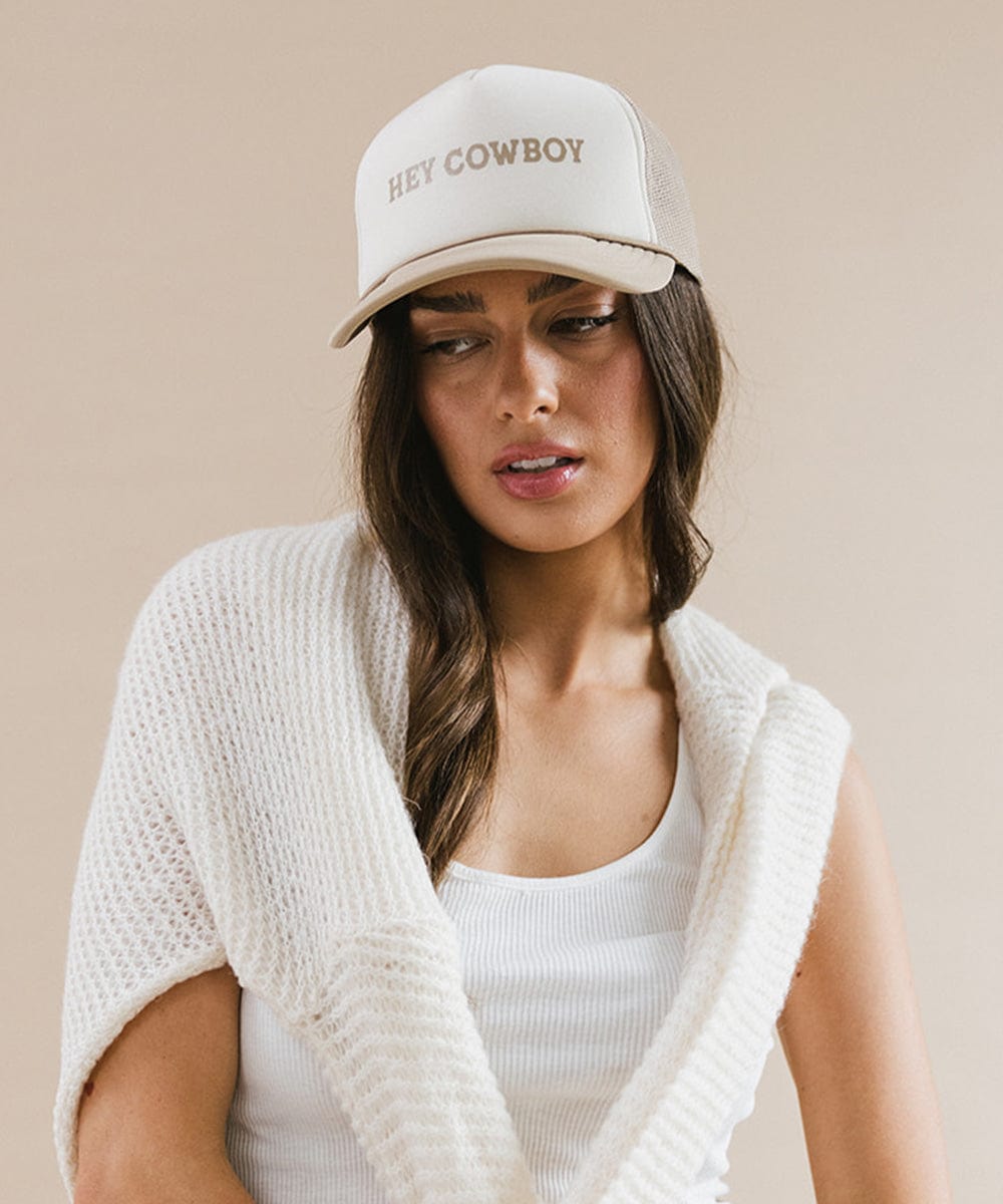 Woman wearing a tan and cream cap with 'Hey Cowboy' text and a white top with a white sweater against a plain background  #color_cream - tan