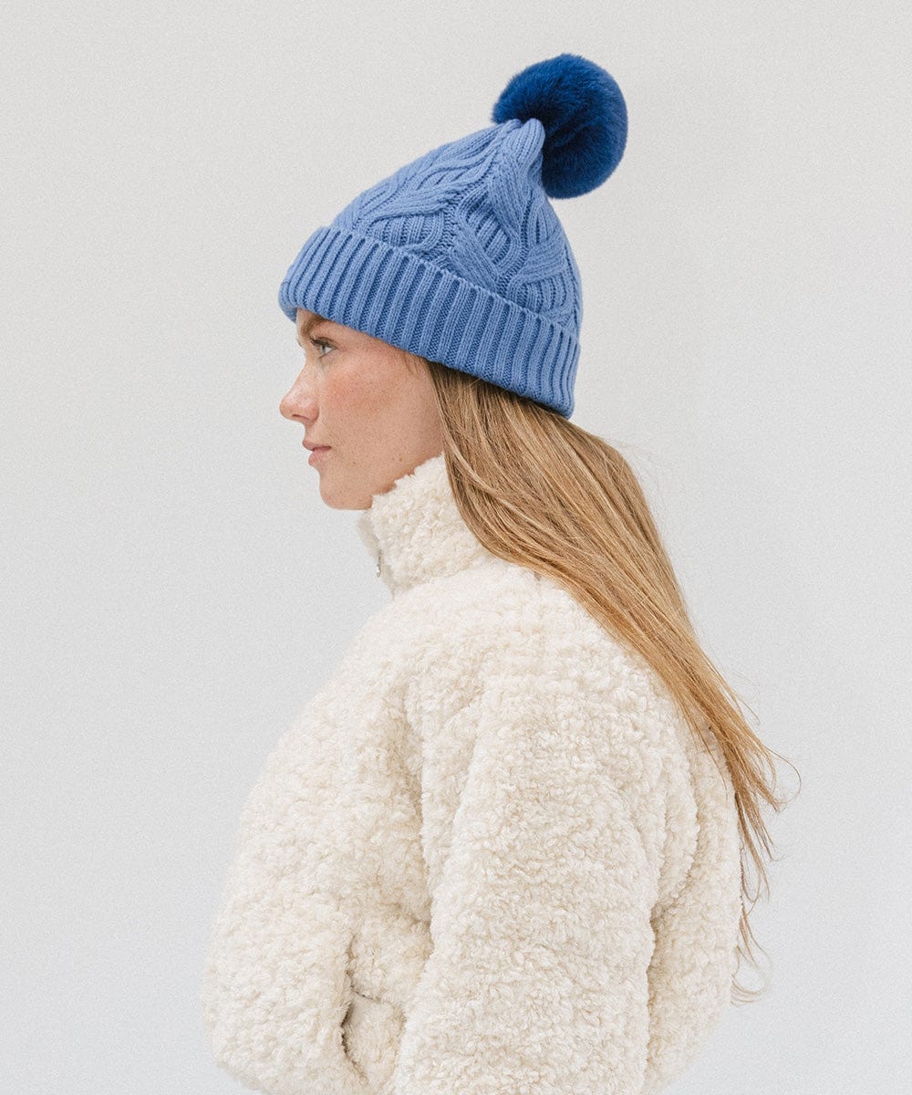 Woman wearing an arctic blue colored knit beanie with a pom-pom and a white sweater, facing sideways against a plain background #color_arctic blue