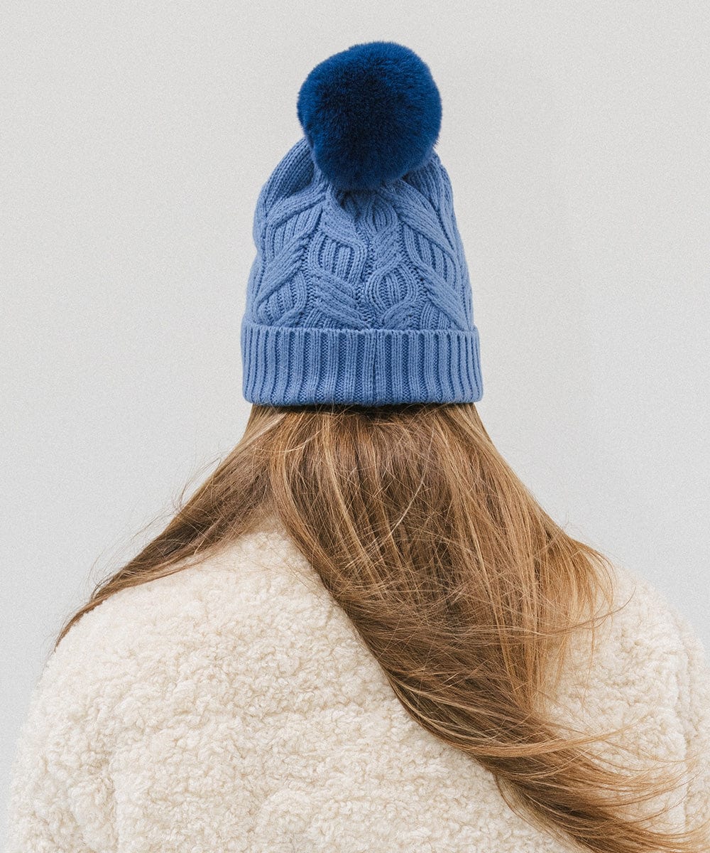 Woman wearing an arctic blue colored knit beanie with a pom-pom and a white sweater, facing behind against a plain background #color_arctic blue