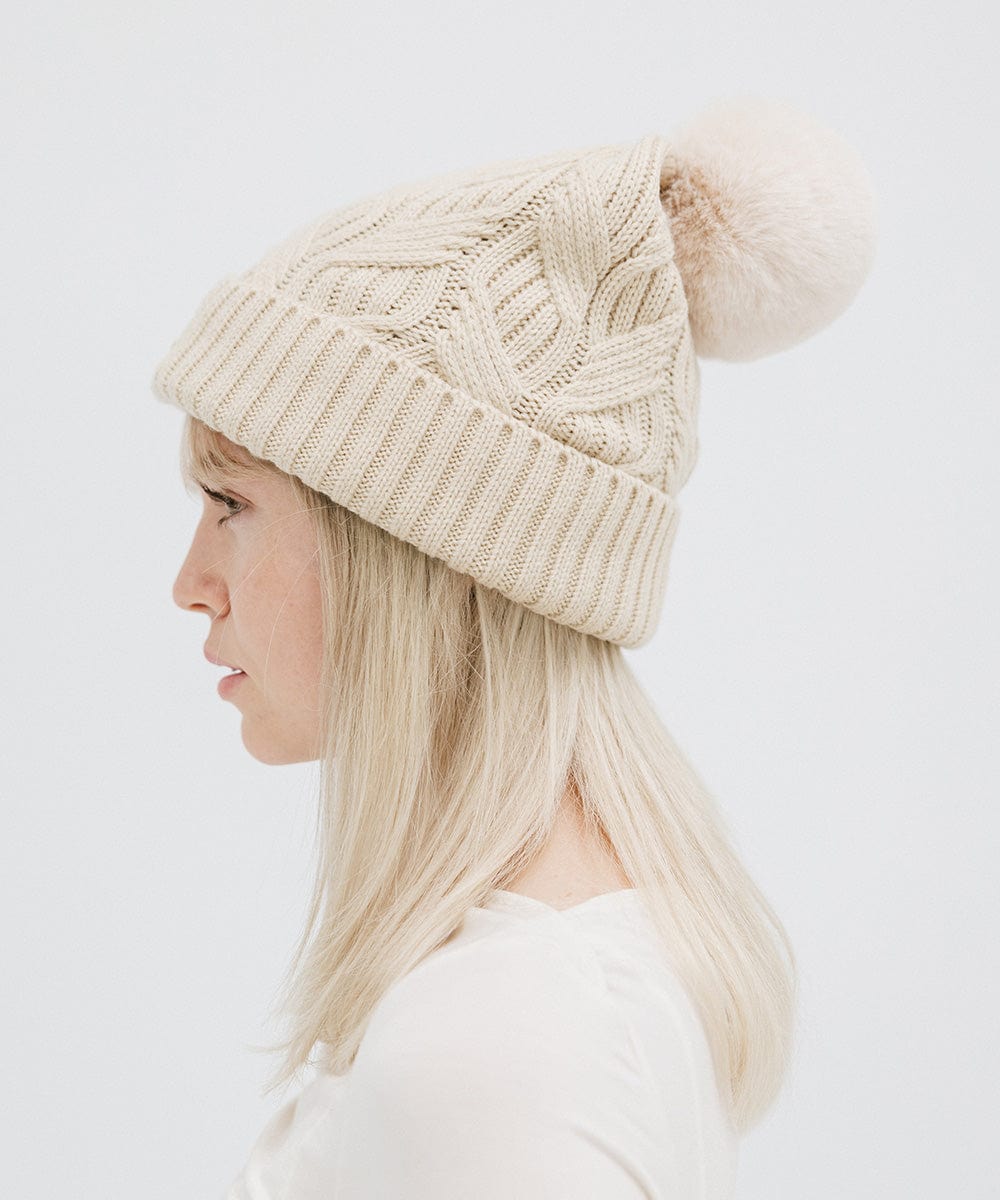 Woman wearing a cream knit beanie with a pom-pom and a white top, facing sideways against a plain background #color_cream