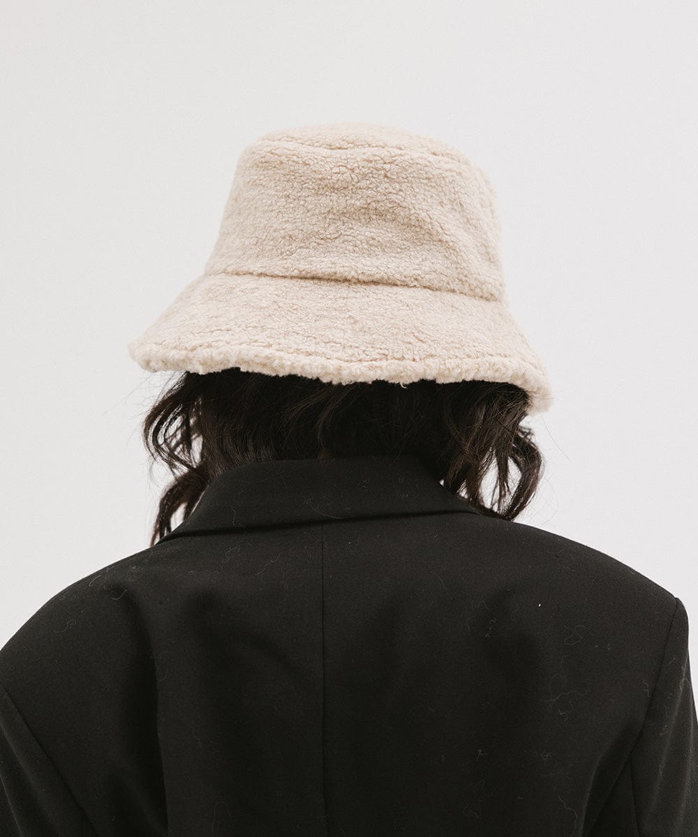 Person wearing a cream sherpa bucket hat and a black coat against a plain background #color_cream