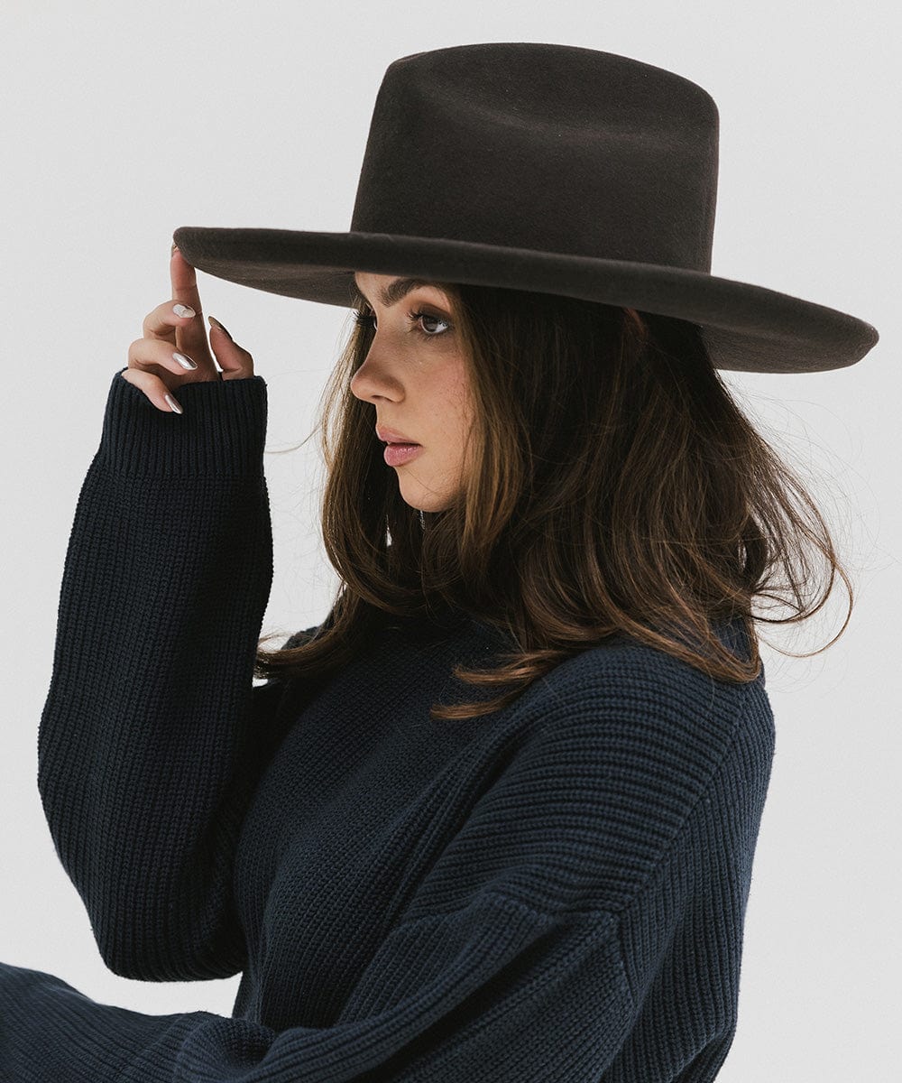 Woman wearing a dark brown western hat with a pencil brim and a tall crown and a dark blue sweater, facing sideways against a plain background  #color_dark brown