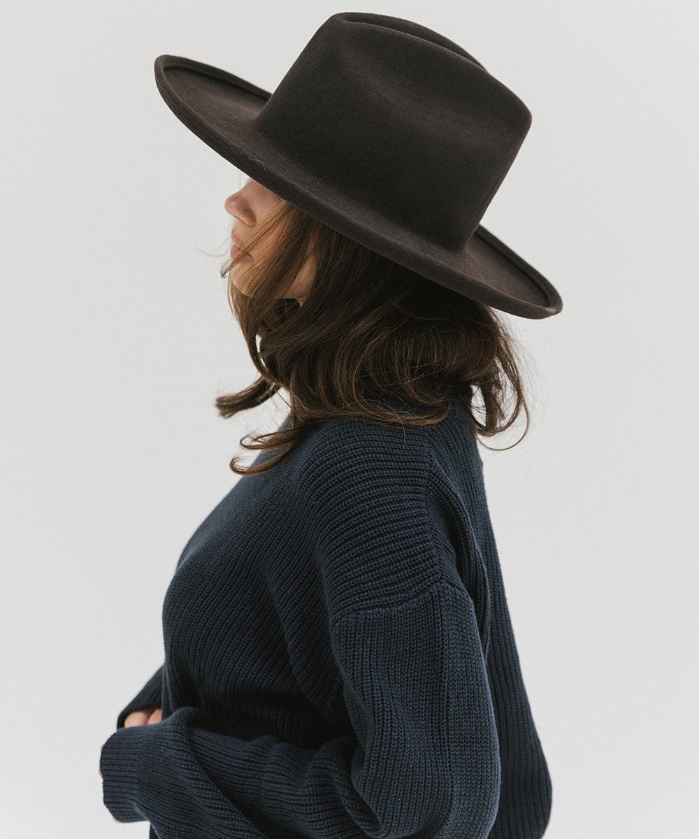 Woman wearing a dark brown western hat with a pencil brim and a tall crown and a dark blue sweater, facing sideways against a plain background  #color_dark brown