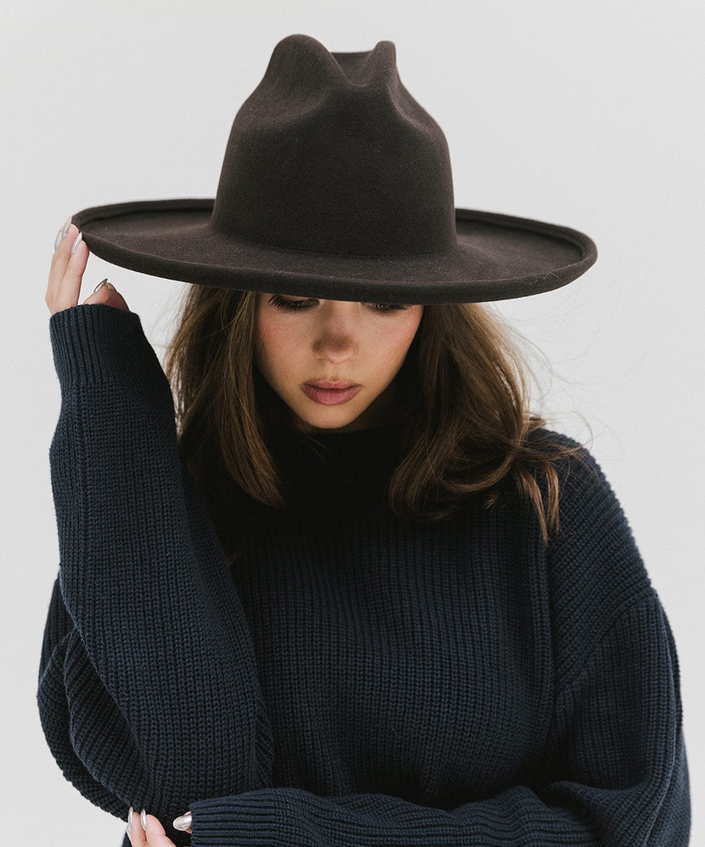 Woman wearing a dark brown western hat with a pencil brim and a tall crown and a dark blue sweater against a plain background  #color_dark brown