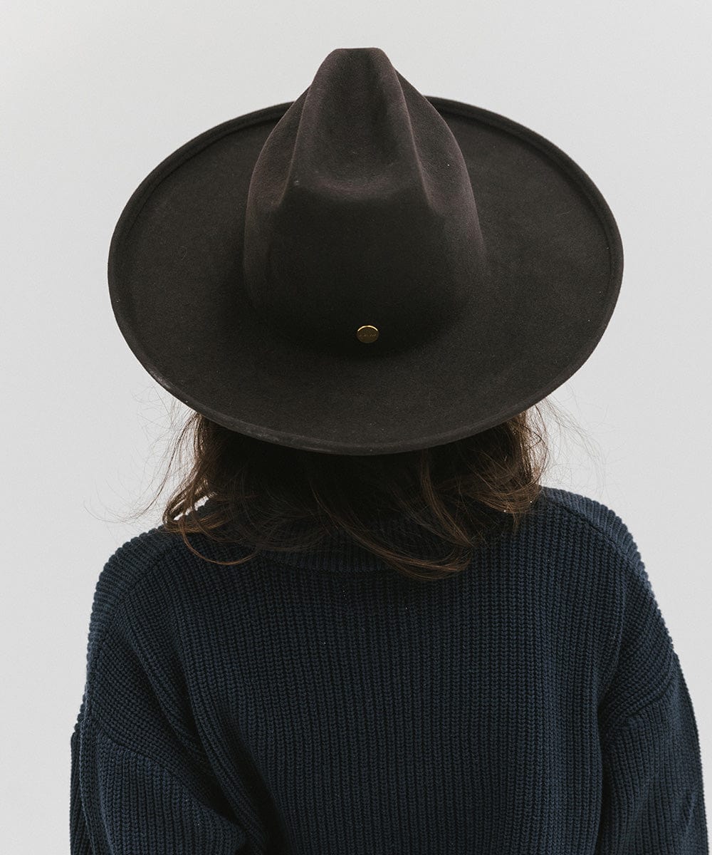 Person wearing a dark brown western hat with a pencil brim and a tall crown and a dark blue sweater against a plain background  #color_dark brown
