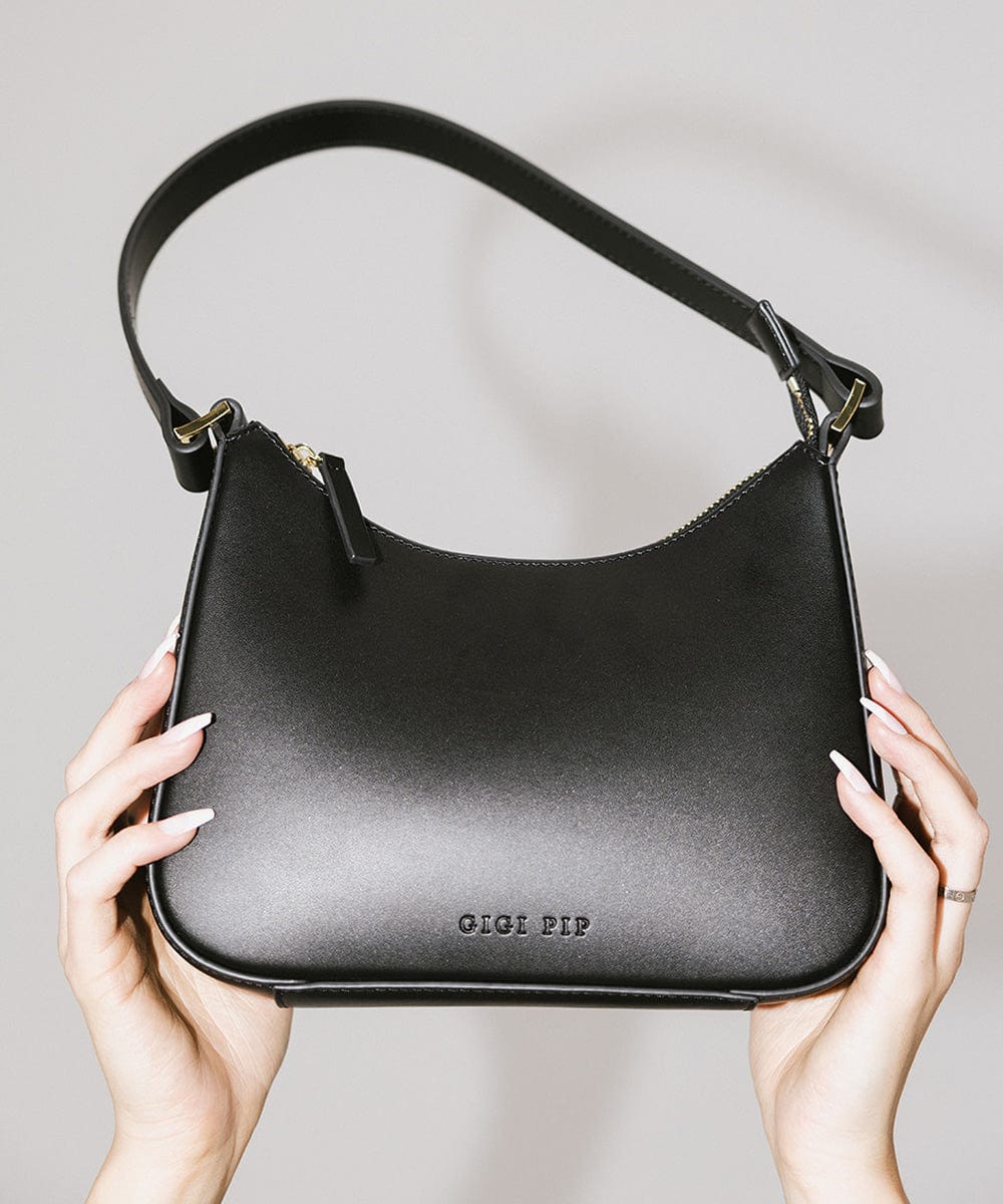 A black genuine leather handbag with a shoulder strap being held by someone against a plain background  #color_black