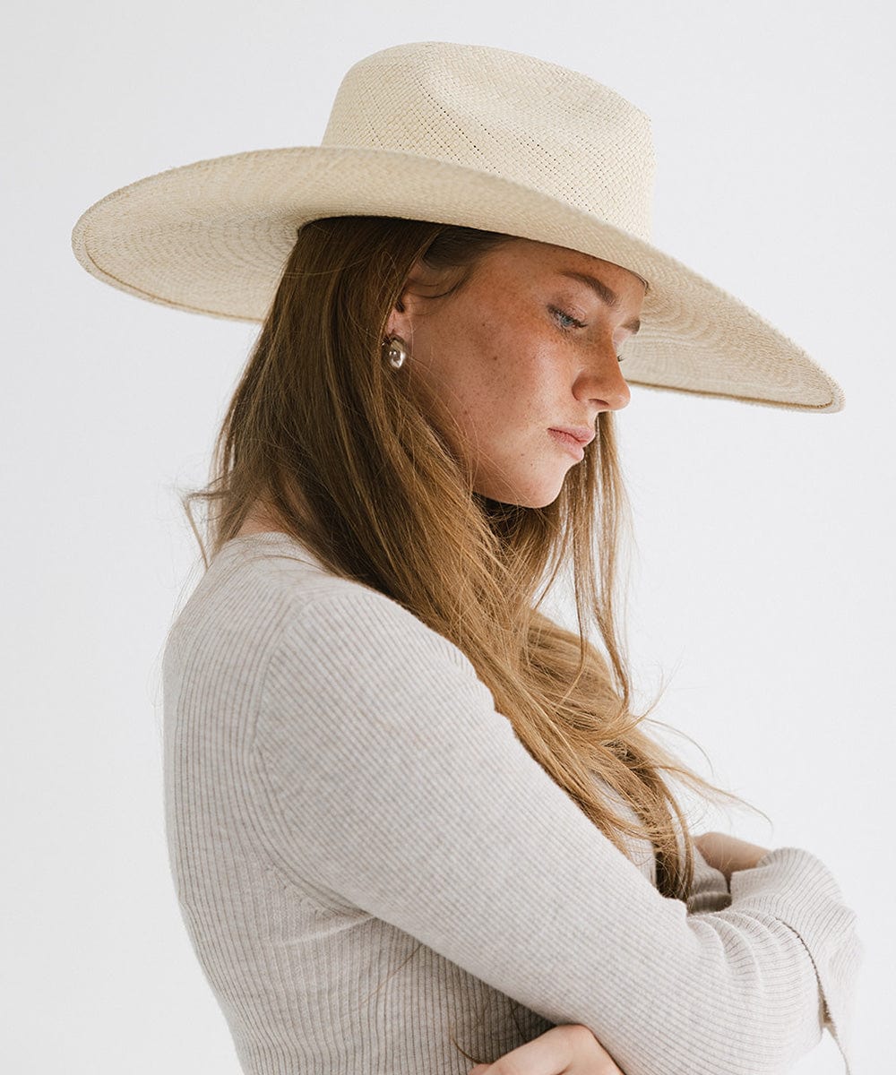 Woman wearing a natural colored wide brim straw hat and a white top, facing sideways against a plain background  #color_natural