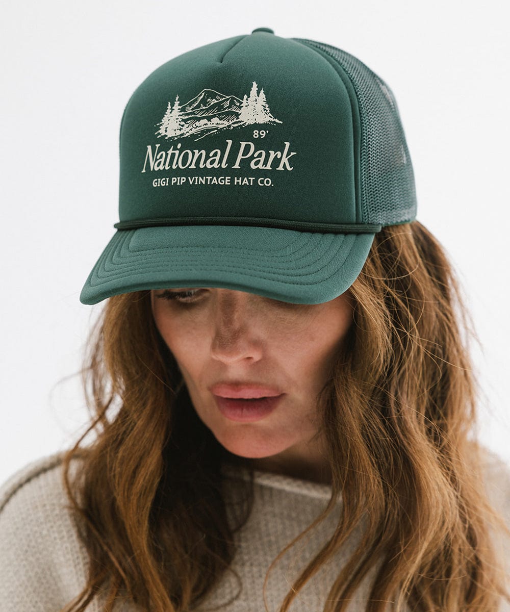 Woman wearing a forest green colored trucker hat with 'National Park' logo against a plain background  #color_forest green