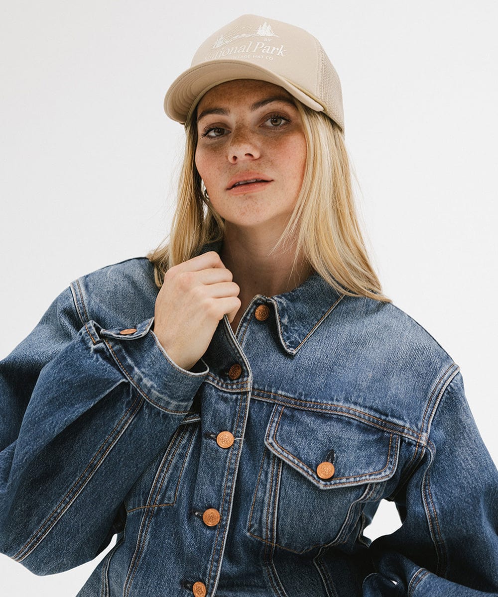 Woman wearing a tan trucker hat with 'National Park' logo and a denim jacket against a plain background  #color_tan