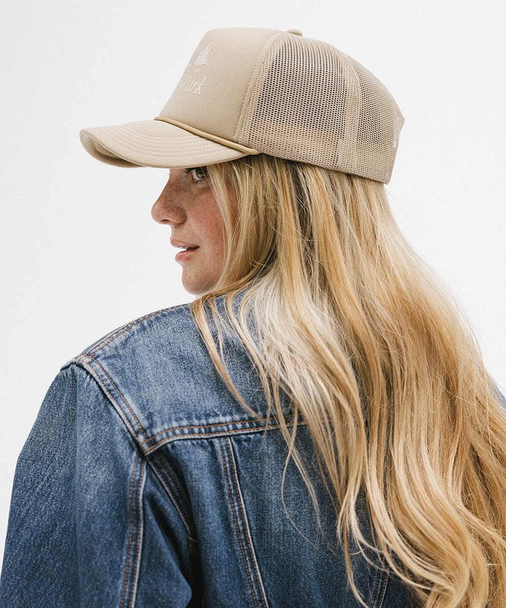 Woman wearing a tan trucker hat with 'National Park' logo and a denim jacket, facing sideways against a plain background  #color_tan