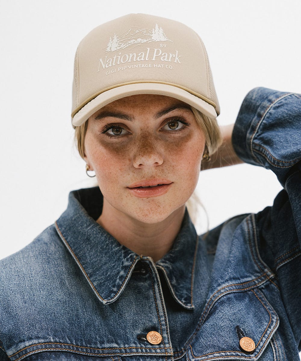 Woman wearing a tan trucker hat with 'National Park' logo and a denim jacket against a plain background  #color_tan