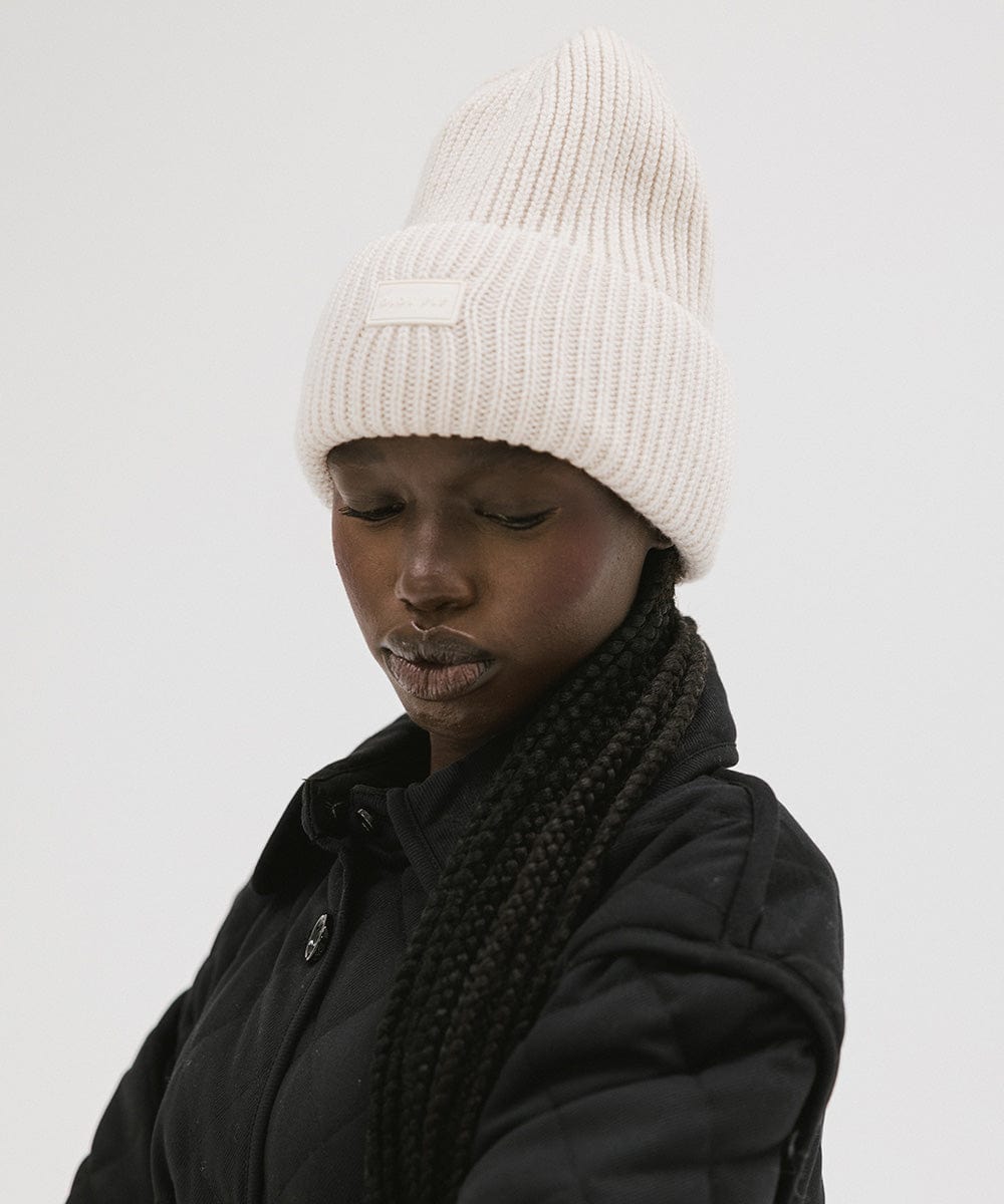 A woman wearing an off white  colored ribbed beanie and a black jacket against a plain background #color_off white
