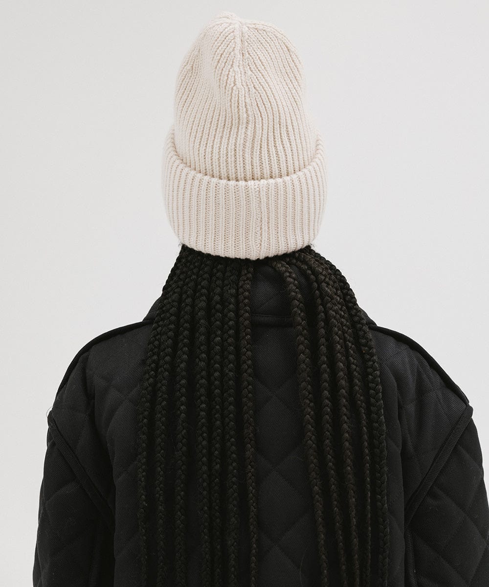 A person wearing an off white  colored ribbed beanie and a black jacket, facing behind against a plain background #color_off white