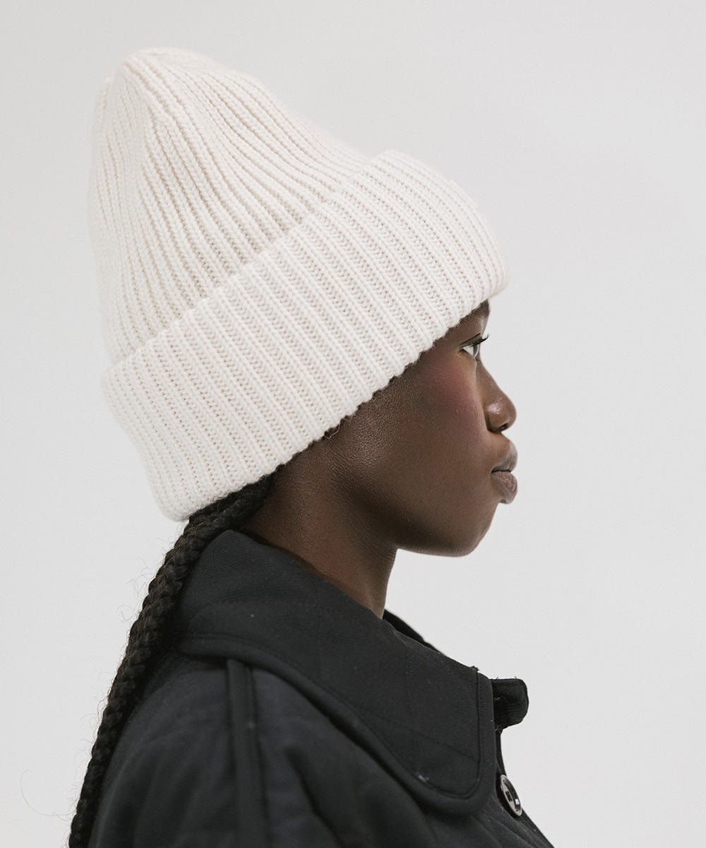 A person wearing an off white  colored ribbed beanie, facing sideways against a plain background #color_off white
