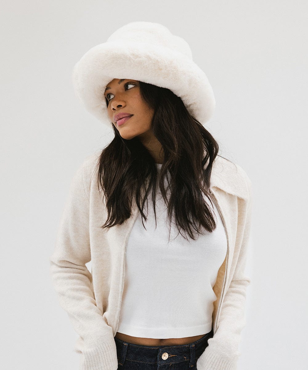Woman wearing a white faux fur bucket hat and a cream jacket against a plain background #color_winter white