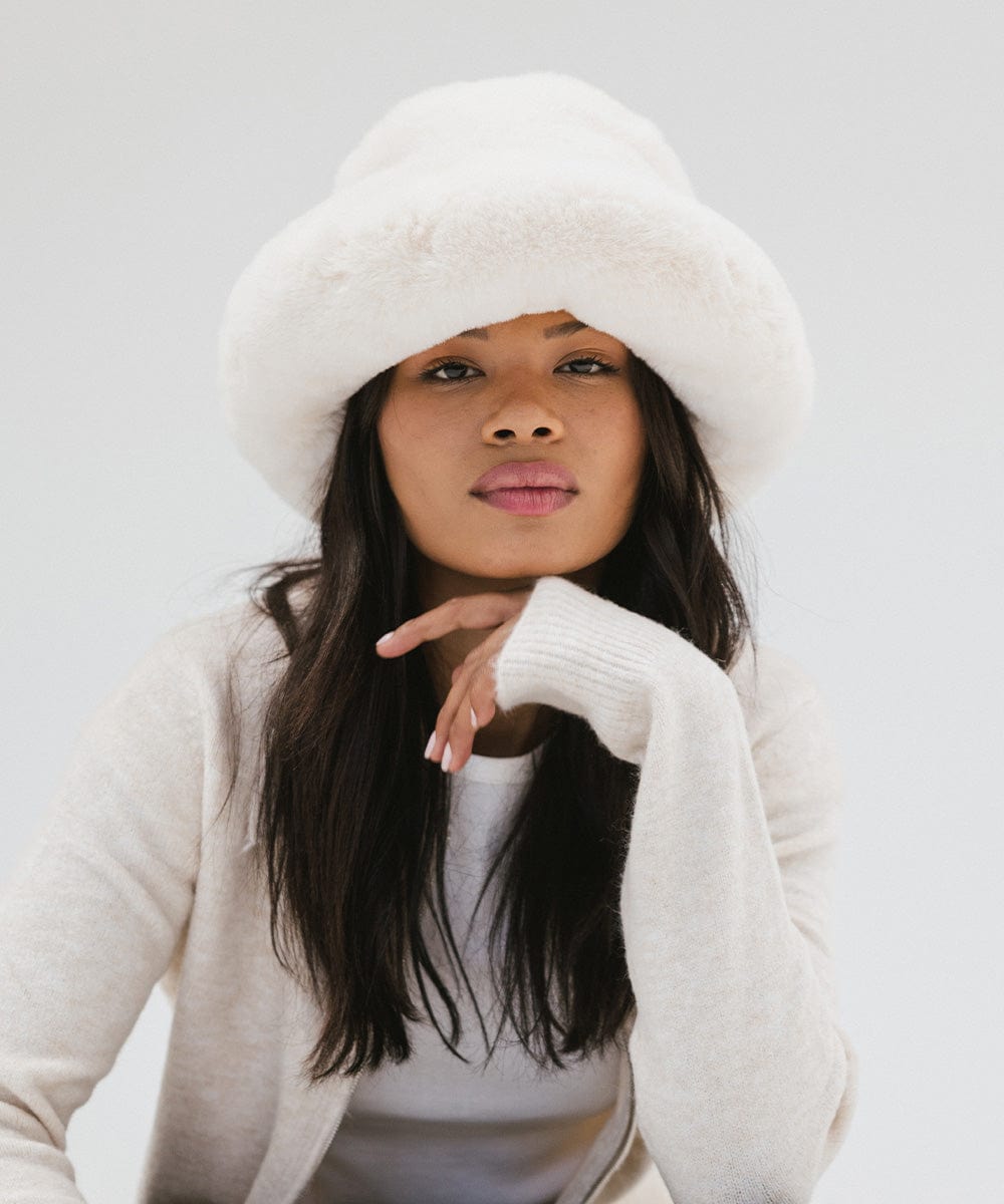 Woman wearing a white faux fur bucket hat and a cream jacket against a plain background #color_winter white