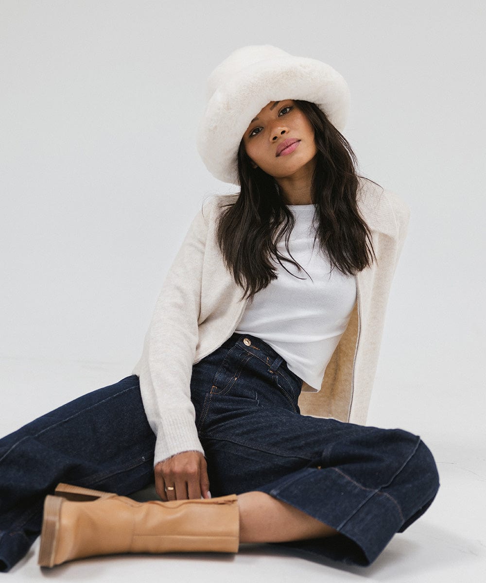 Woman wearing a white faux fur bucket hat, a cream jacket, blue jeans, and brown boots, sitting against a plain background #color_winter white