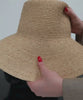 A natural colored oversized packable straw bucket hat being folded and packed into a black bag against a plain background #color_natural