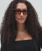 Woman wearing sunglasses with black frame and orange lenses and a black t-shirt with a black shirt, posing against a plain background #color_black