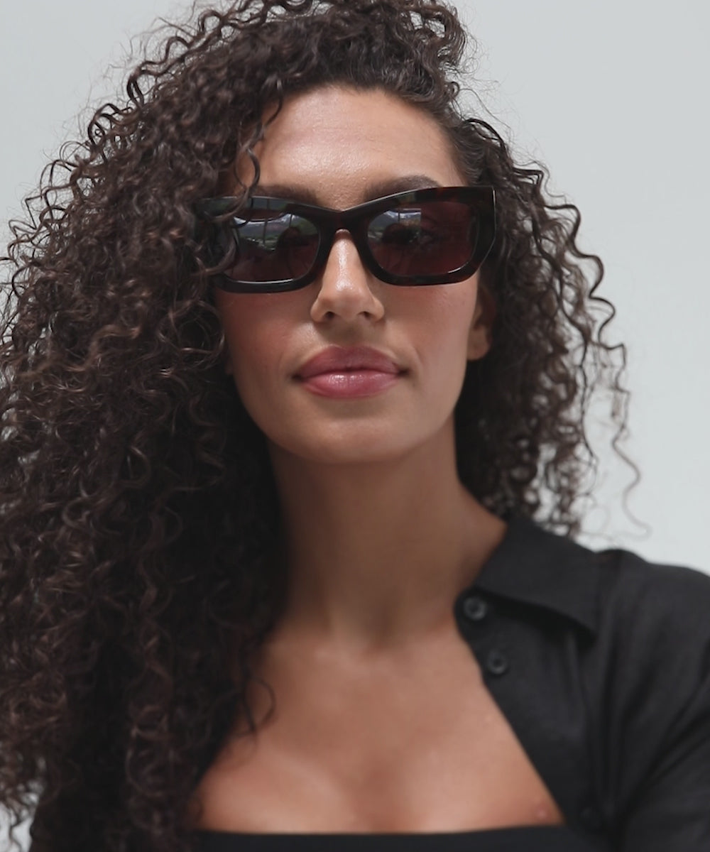 Woman wearing tortoise pattern sunglasses and a black shirt, posing against a plain background  #color_tortoise
