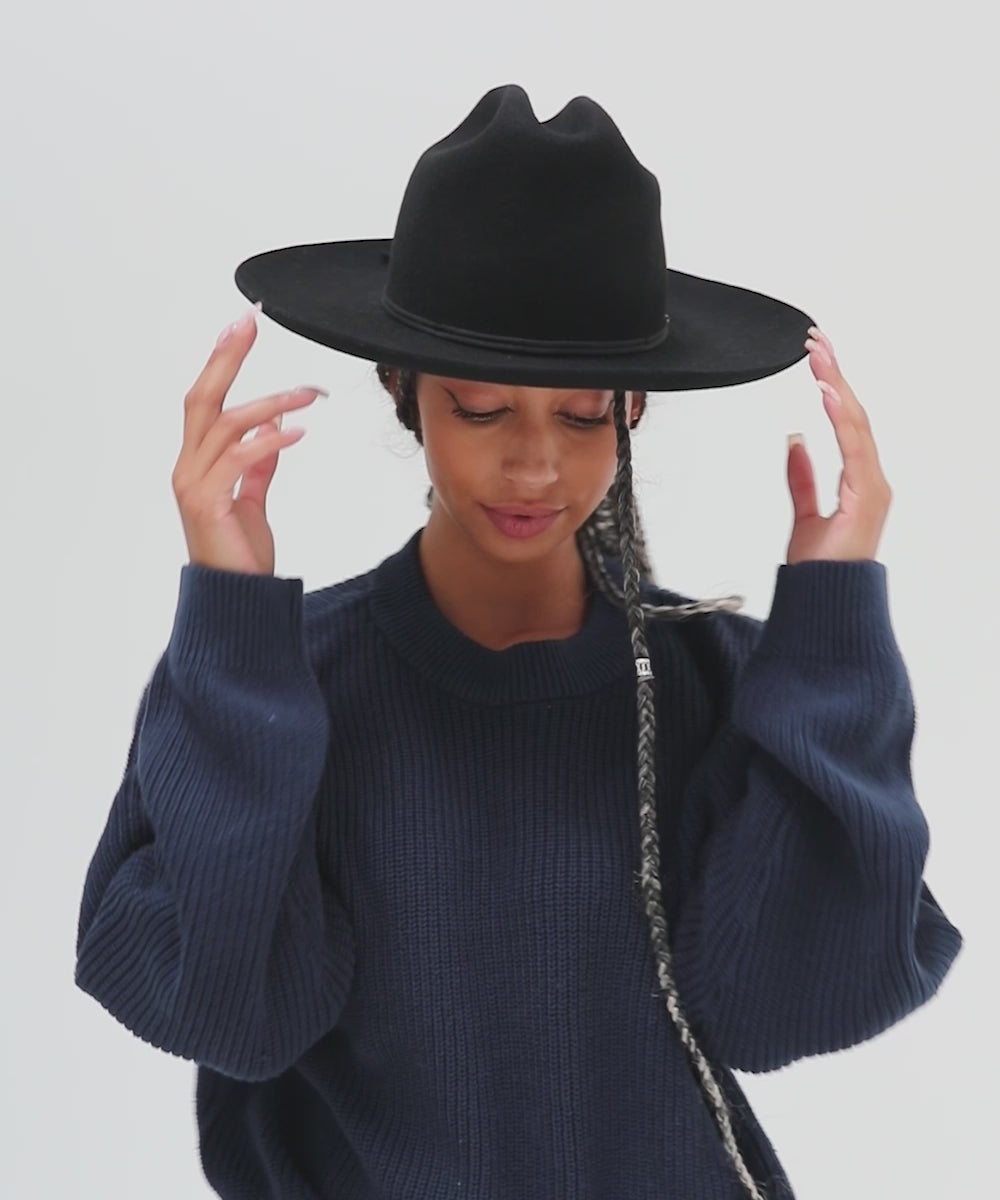 Woman wearing a black Western hat with a classic cattleman crown and a black hat band, and a dark blue sweatshirt, posing against a plain background  #color_black