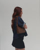 Woman wearing a denim shirt and a denim skirt, carrying a chocolate colored genuine leather handbag against a plain background #color_chocolate