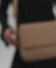 Woman wearing a black outfit carrying a tan Rhys Crossbody Genuine Leather Bag, posing against a plain background #color_tan