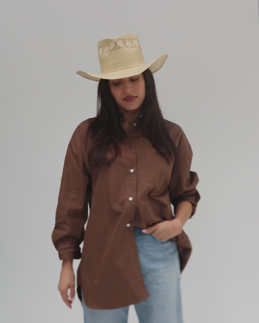 Woman wearing a natural colored straw hat, a brown shirt and blue jeans, posing against a plain background #color_natural 