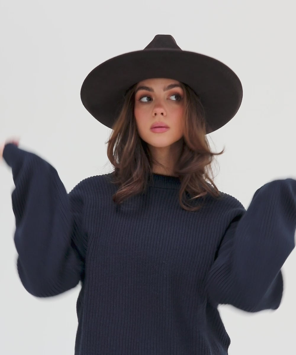 Woman wearing a dark brown western hat with a pencil brim and a tall crown with a hat band, and a dark blue sweater against a plain background  #color_dark brown