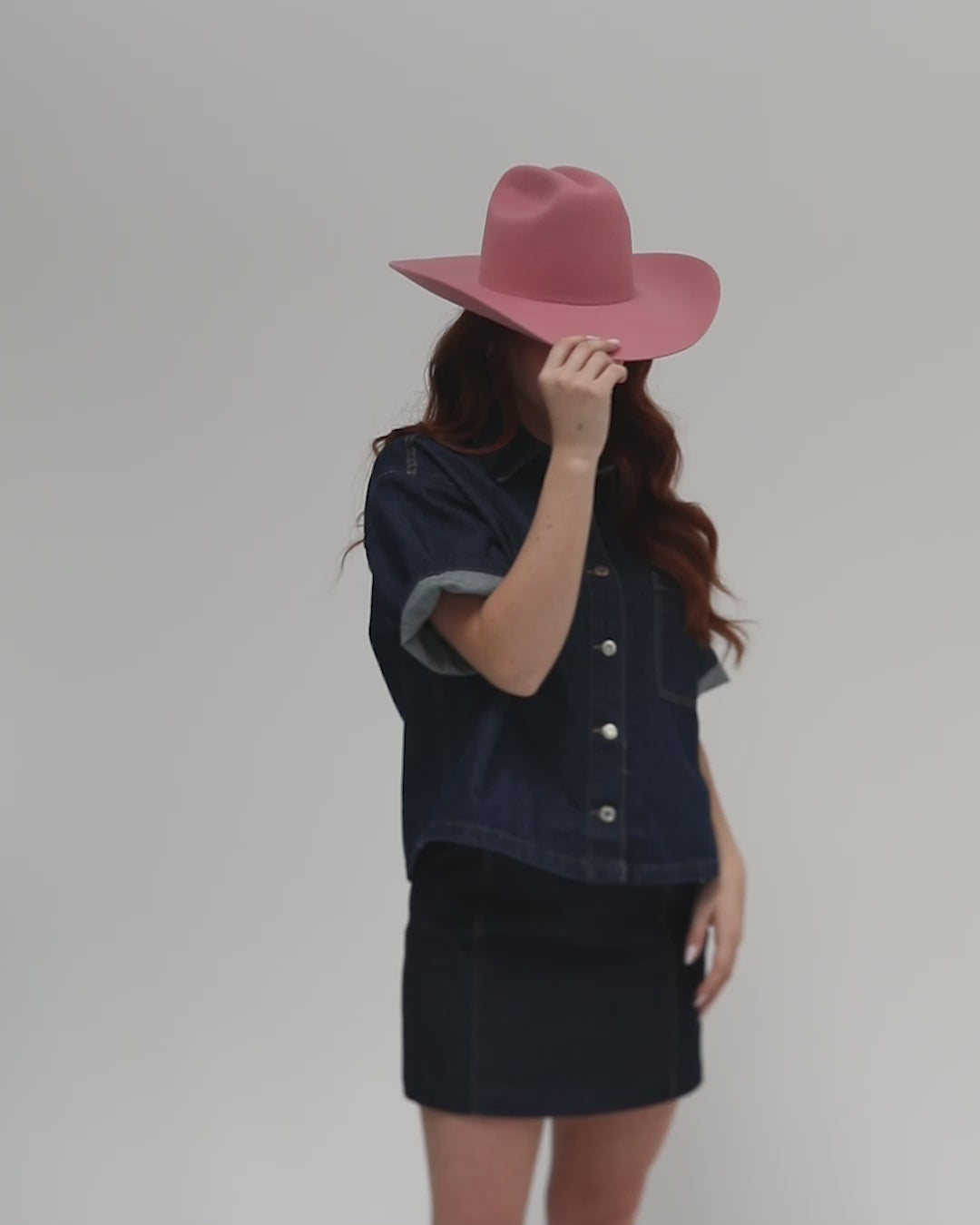 Woman wearing a rose colored cowboy hat, a dark blue denim shirt and a dark blue denim skirt, posing against against a plain background #color_rose