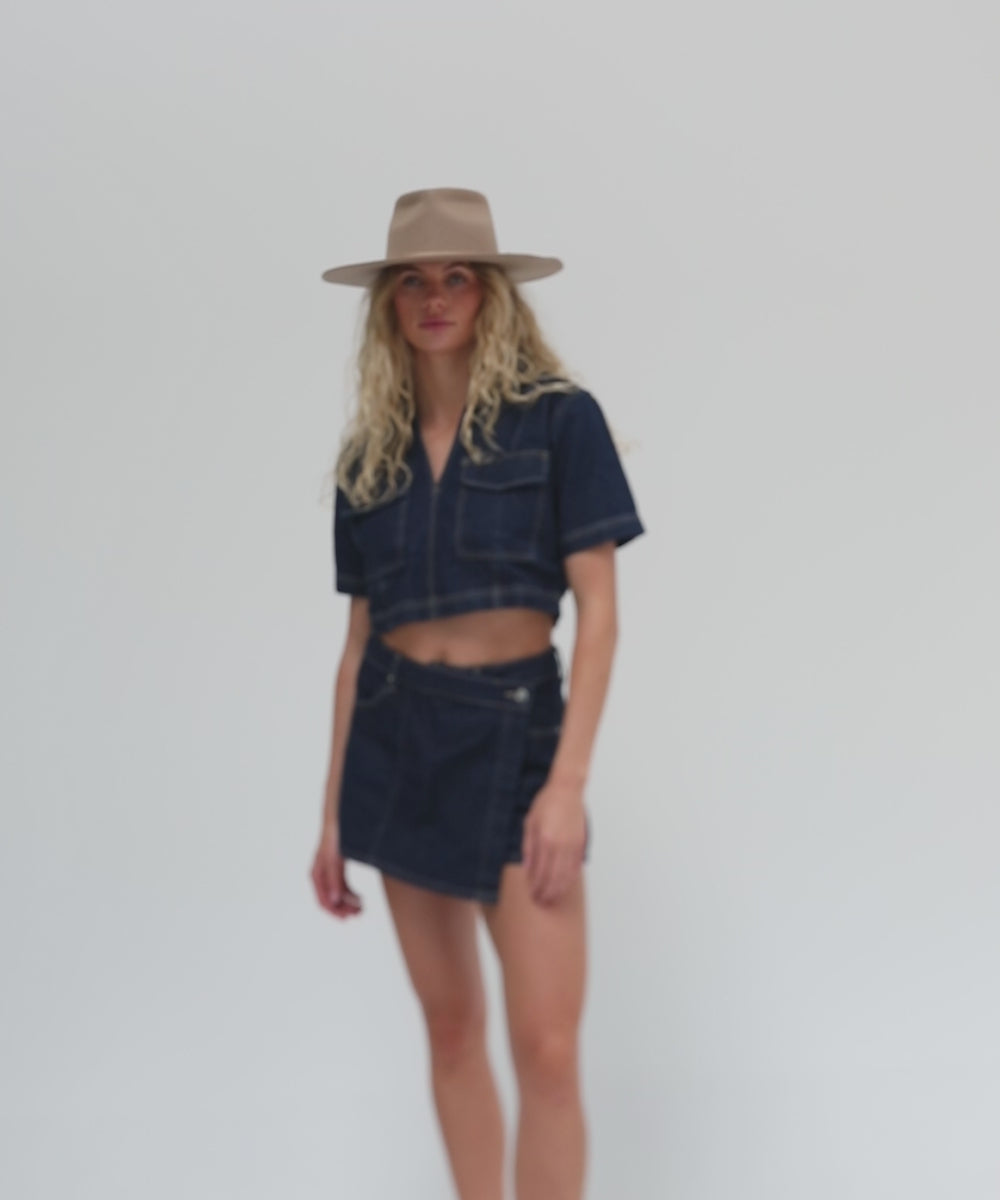 Woman with long blonde hair wearing a tan fedora hat, a denim top and a denim skirt, posing against a plain background  #color_tan