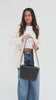 Woman wearing a white top with a cream coat and blue jeans, carrying a black Rhys Crossbody Genuine Leather Bag, posing sideways against a plain background #color_black