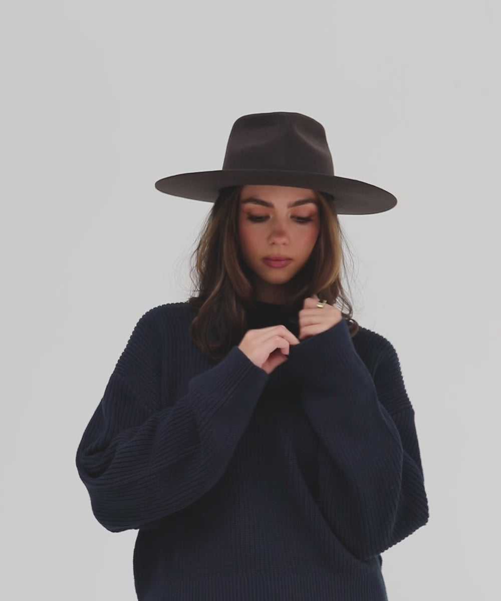 Woman wearing a dark brown fedora hat with a hat band and a dark blue sweatshirt, posing against a plain background #color_dark brown