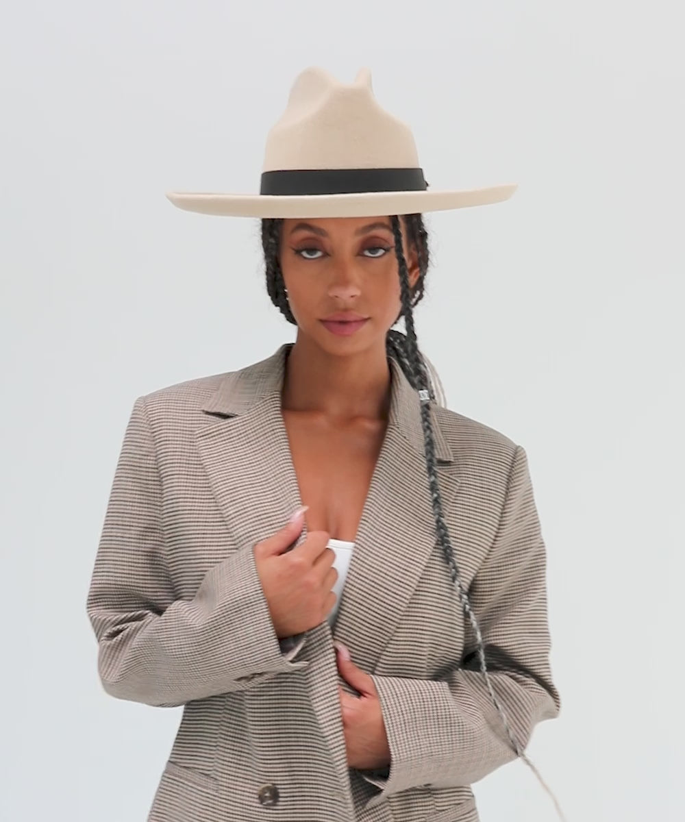 Woman wearing an off white western hat with a pencil brim and a tall crown and a white top with a coat against a plain background #color_off white