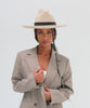 Woman wearing an off white western hat with a pencil brim and a tall crown and a white top with a coat against a plain background #color_off white