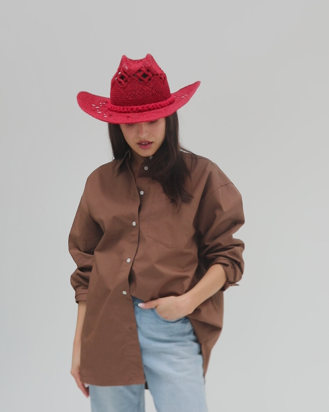 Woman wearing a cherry red colored paper rope straw cowboy hat, a brown shirt and blue jeans, posing against a plain background #color_cherry red
