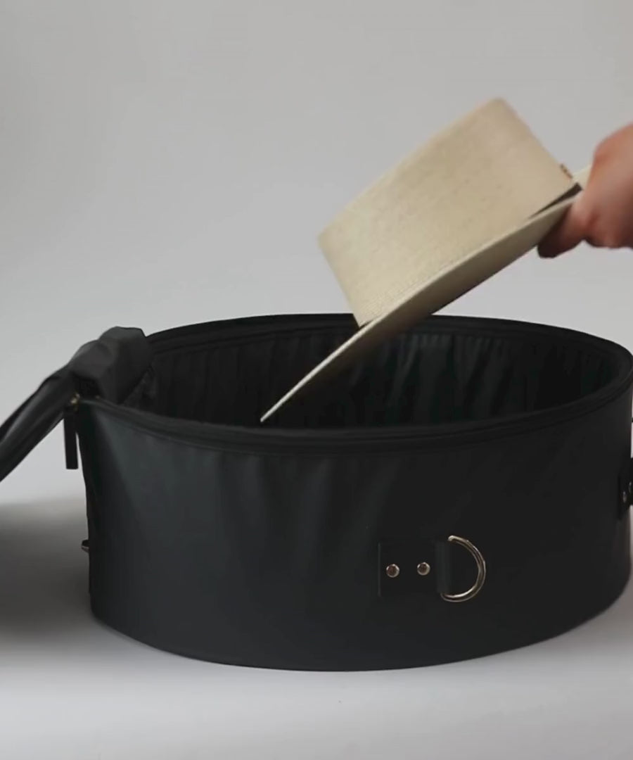 Hats being packed in a round black hat travel case and being carried by a woman wearing a grey outfit #color_black