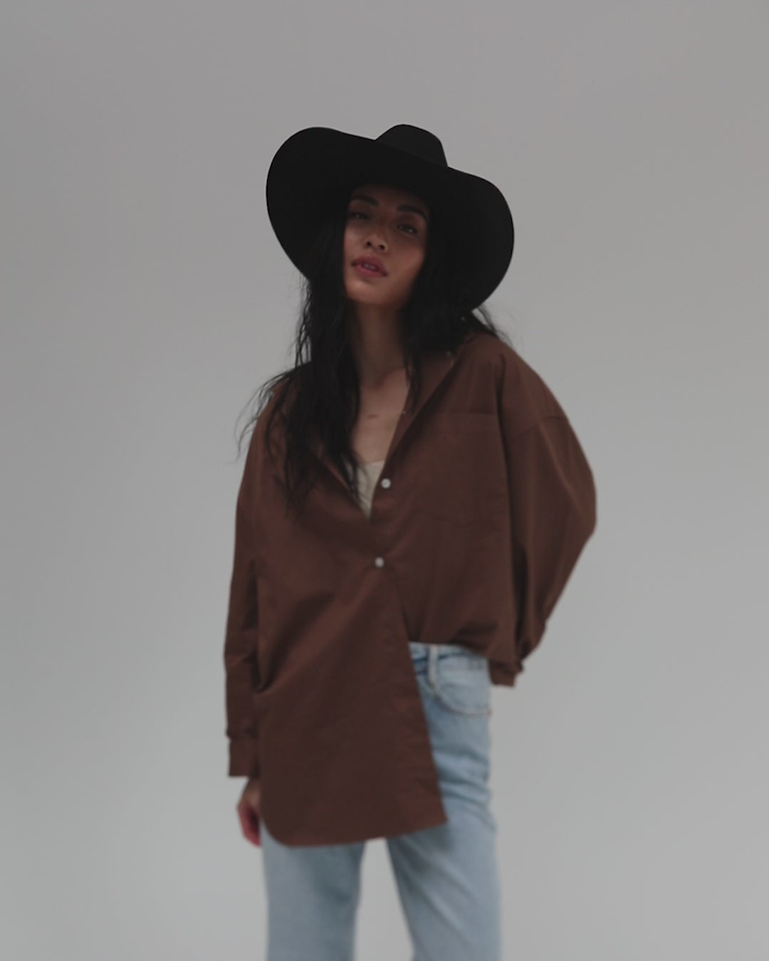 Woman wearing a black cowboy hat, a brown shirt and light blue jeans, posing against a plain background #color_black