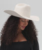 Woman wearing an off white cowboy hat and a black top with a black shirt, posing against a plain background
#color_off white