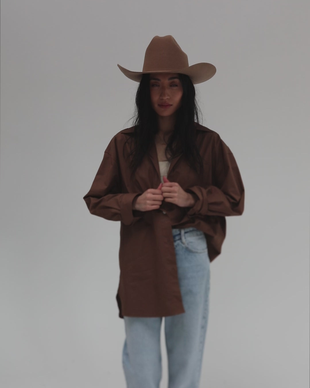 Woman wearing a brown cowboy hat, a brown shirt and light blue jeans, posing against a plain background #color_brown