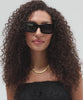 Woman wearing black sunglasses, a gold chain and a black top, posing against a plain background  #color_black
