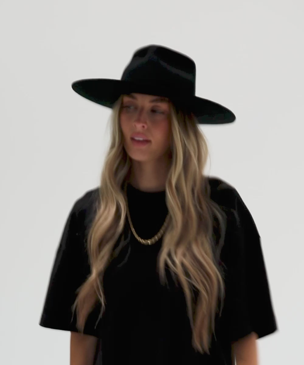 Woman with long hair wearing a black Emery Teardrop Fedora Hat with a hat band, a gold chain and a black t-shirt, posing against a plain background #color_black