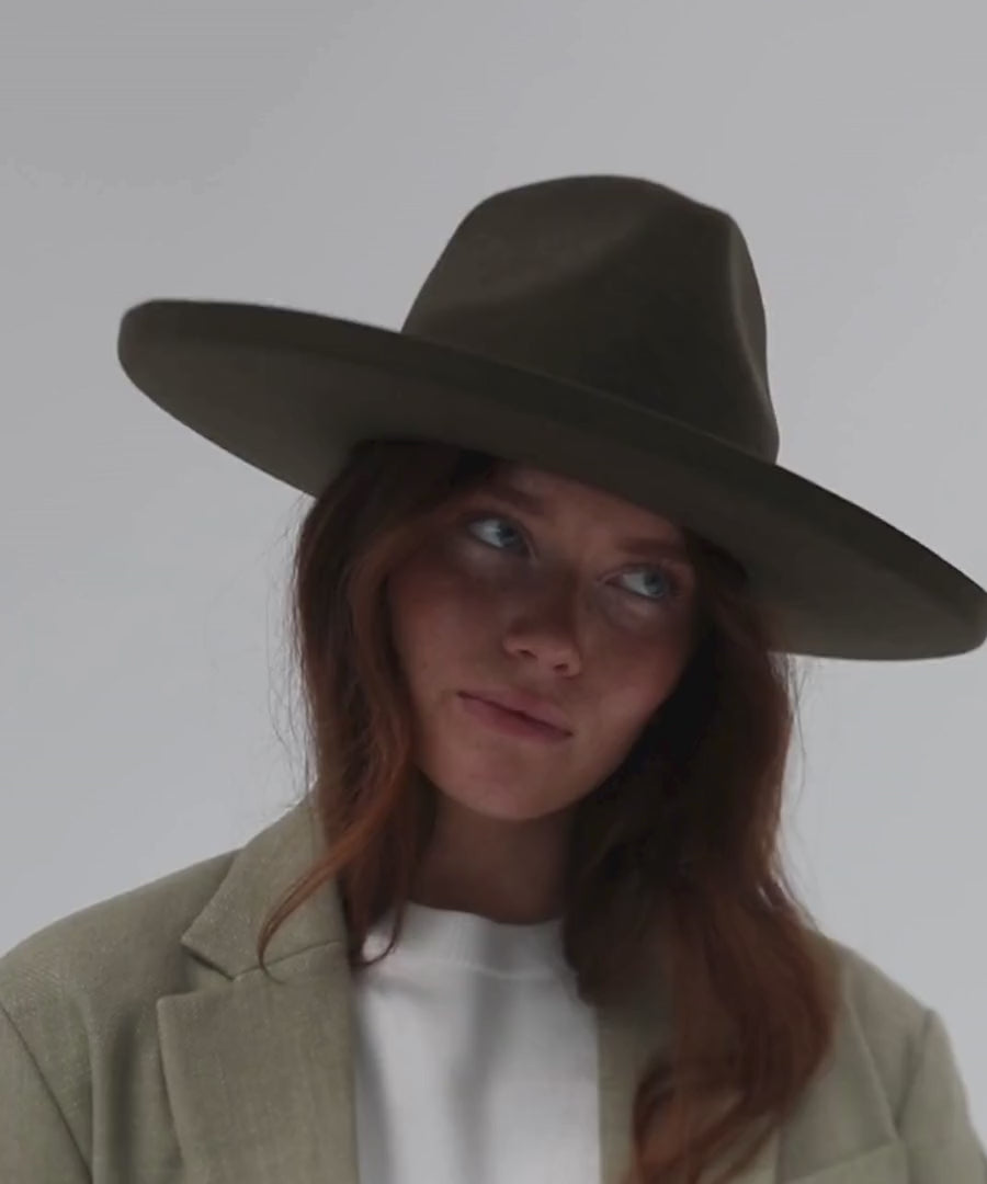 Woman wearing a moss colored pencil brim fedora hat with a wide brim and a white t-shirt with a beige blazer, posing against a plain background #color_limited edition moss