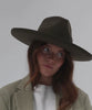Woman wearing a moss colored pencil brim fedora hat with a wide brim and a white t-shirt with a beige blazer, posing against a plain background #color_limited edition moss
