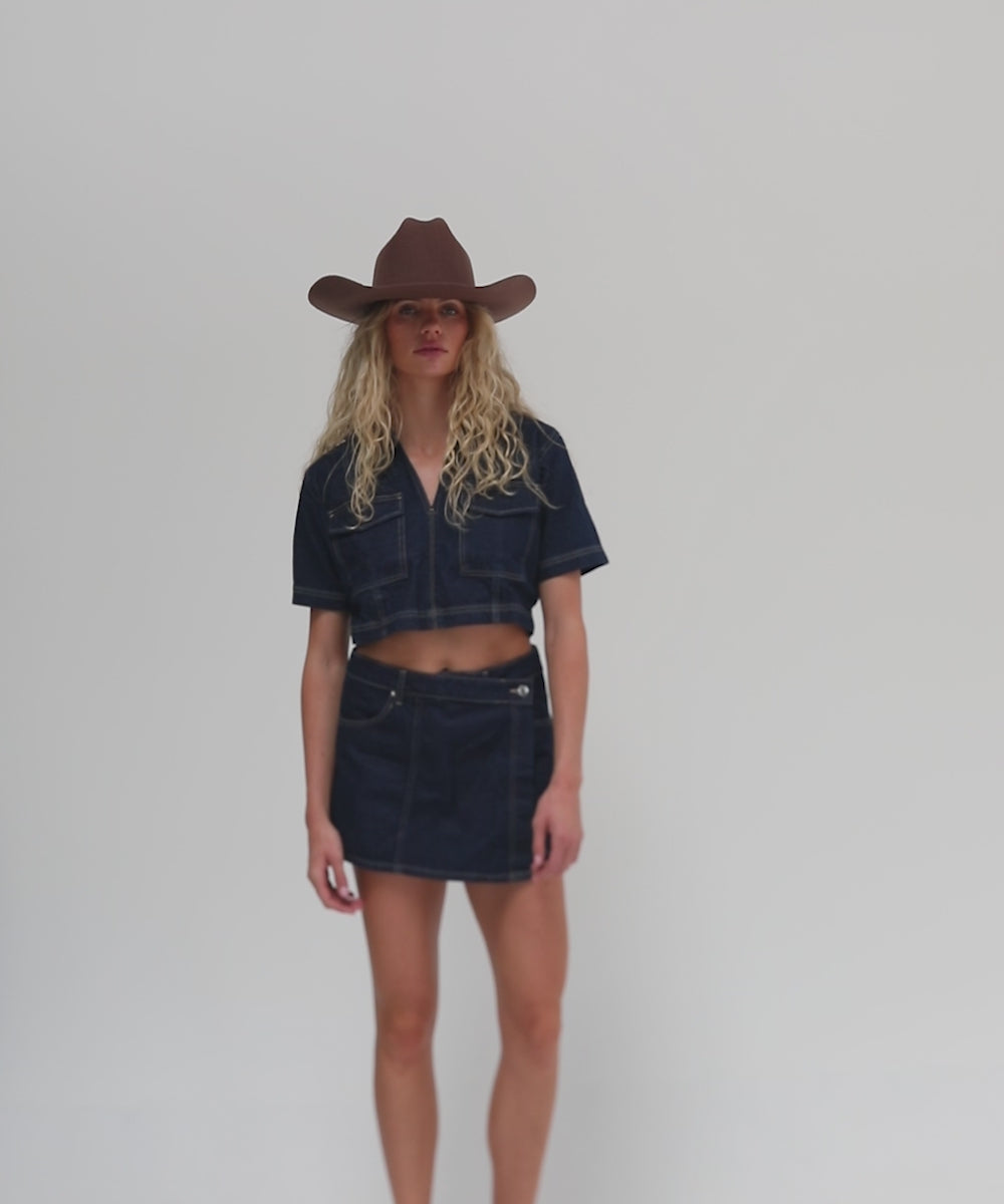 Woman wearing a chocolate colored cowboy hat, a dark blue denim shirt and a dark blue denim skirt, posing against a plain background #color_chocolate