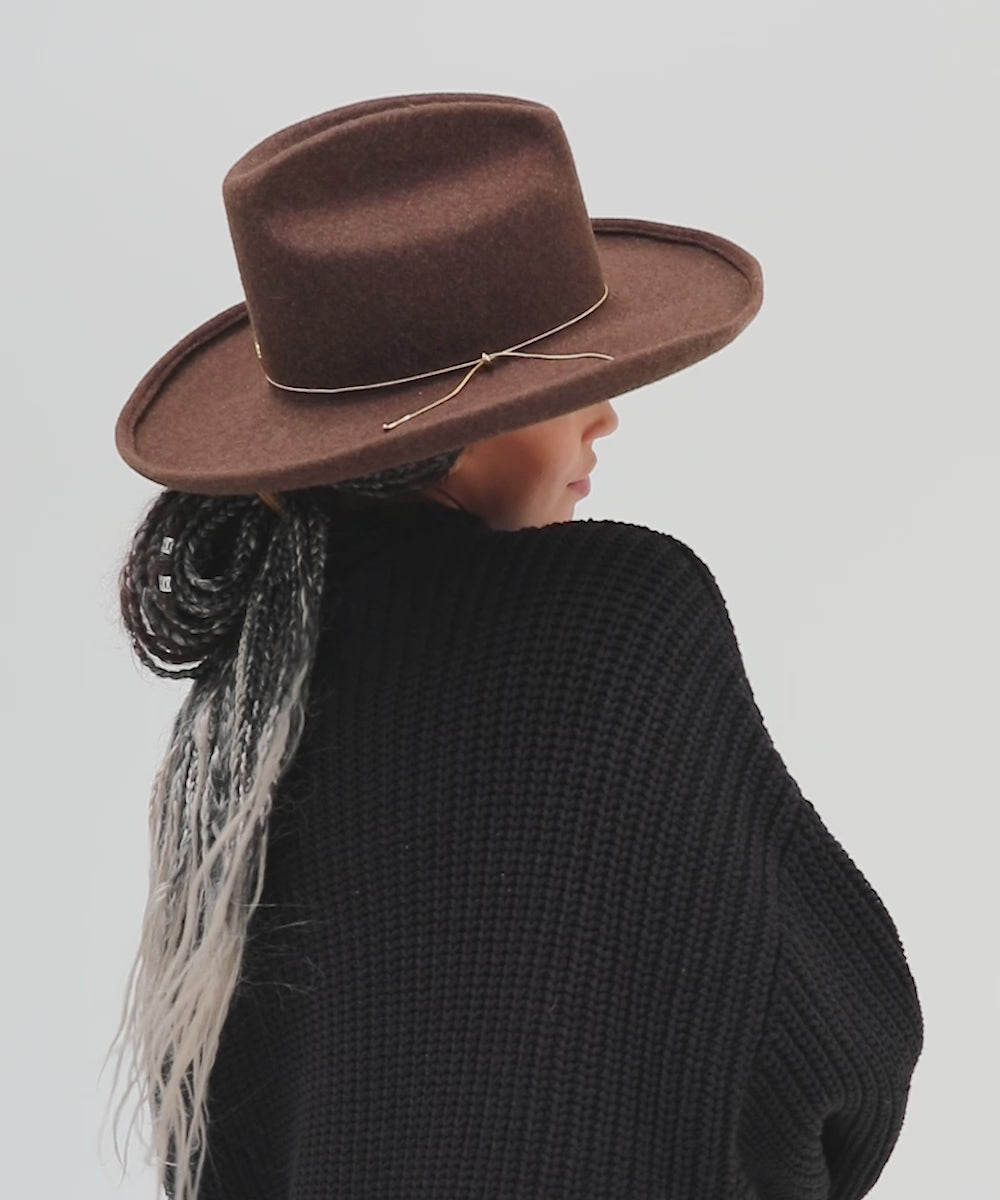 Woman wearing a mix brown colored western hat with a pencil brim and a tall crown with a hat band, and a black sweater against a plain background  #color_mix brown