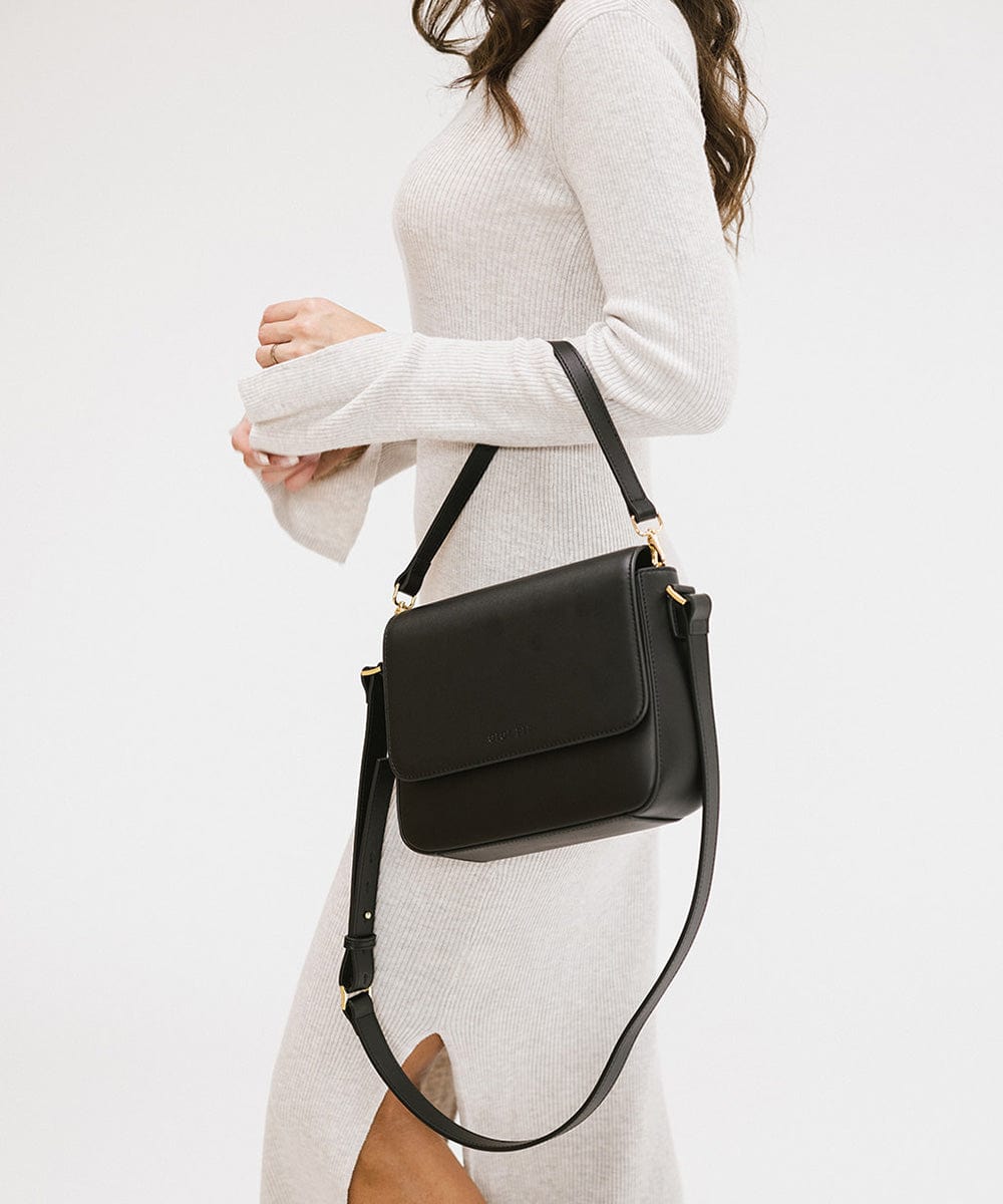 Person wearing a white outfit carrying a black Rhys Crossbody Genuine Leather Bag, posing sideways against a plain background  #color_black
