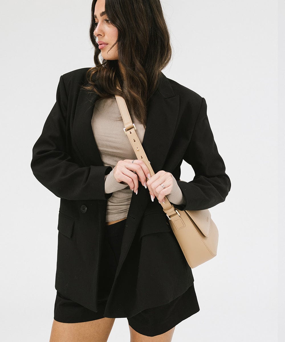 Woman wearing a black blazer and black shorts, carrying a tan Rhys Crossbody Genuine Leather Bag against a plain background  #color_tan