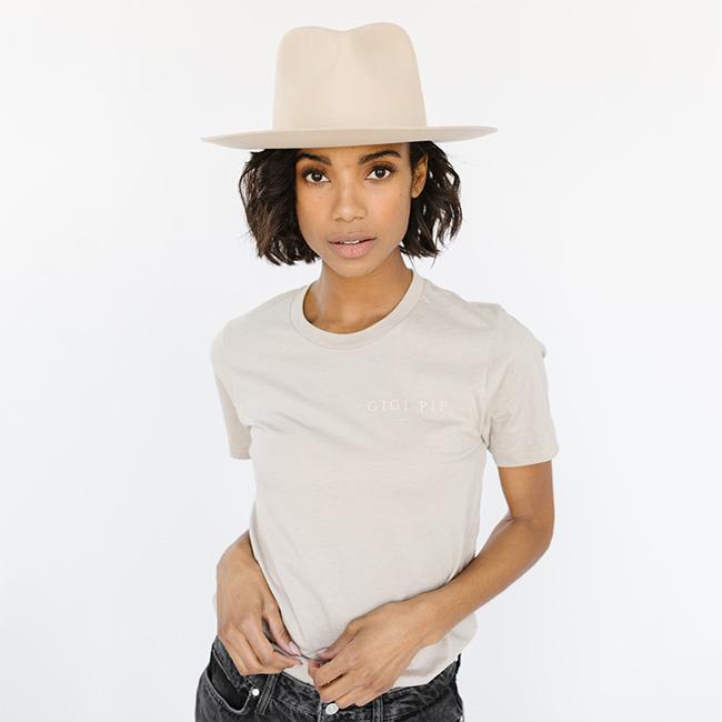 Woman wearing a sand colored t-shirt with 'GIGI PIP' text and a cream hat against a white background #color_sand