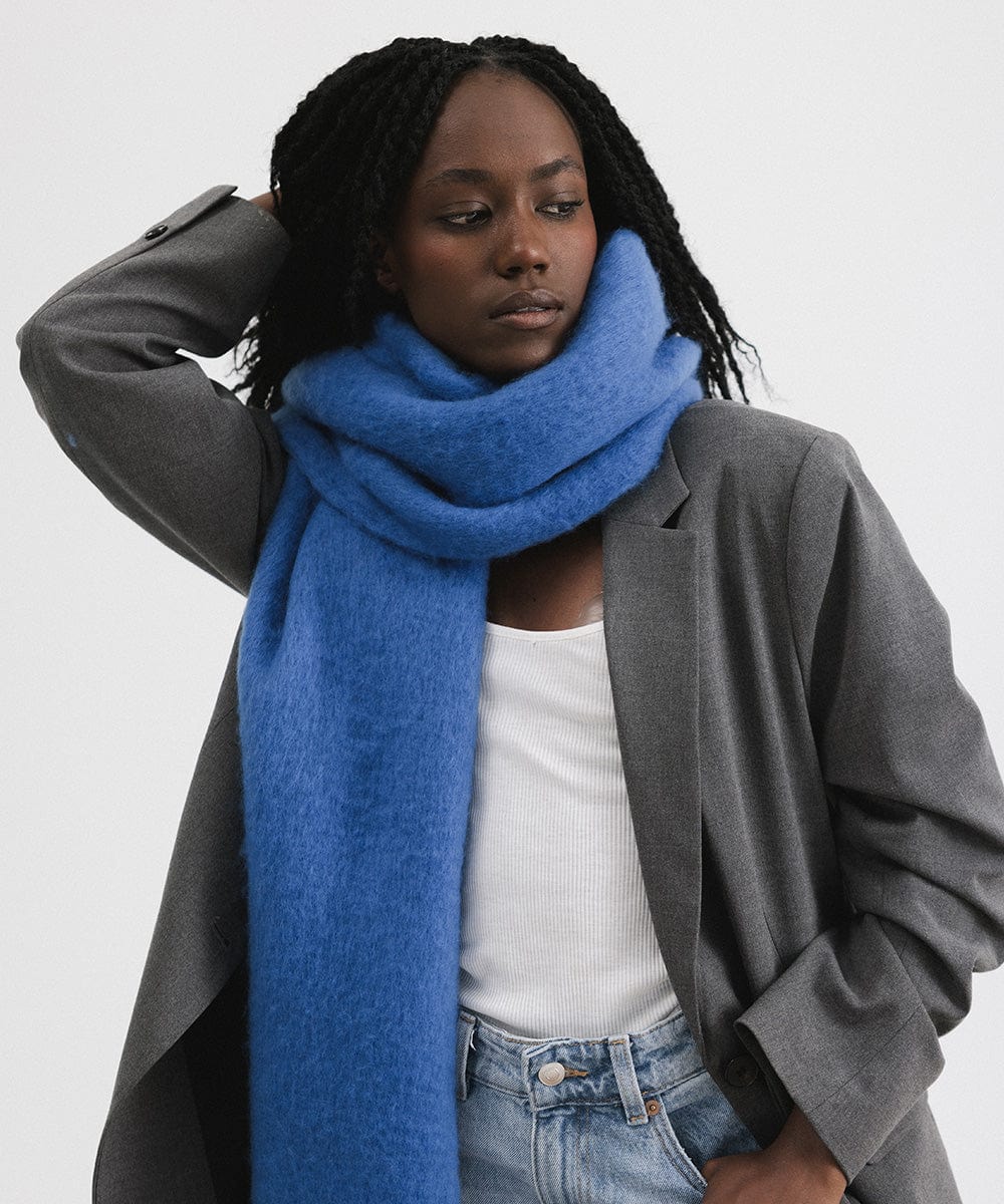 A woman wearing a grey blazer and an alpine blue colored oversized knit scarf with fringes, draped over the neck against a plain background  #color_alpine blue